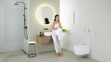 Жінка спирається на умивальник у ванній кімнаті з унітазом-біде Geberit AquaClean Sela й умивальником і меблями Geberit VariForm