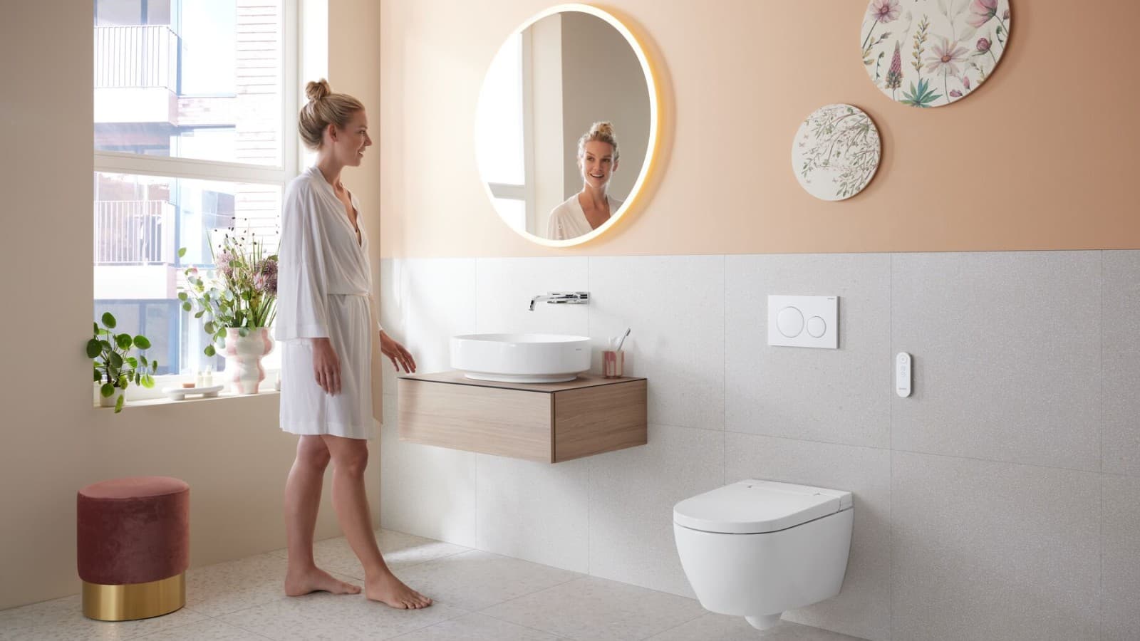 Жінка дивиться в дзеркало у ванній кімнаті з унітазом-біде AquaClean Alba Жінка дивиться в дзеркало у ванній кімнаті з унітазом-біде AquaClean Alba